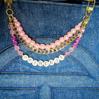 A close-up of a double-layered beaded bag charm draped over a blue denim tote bag. The bag charm features a top layer of light pink pearls and a bottom layer of gold curb linked chains, accented with faceted pink and deep pink crystals and a white round letter bead spelling 'PRINCESS'. All hardware is gold including the lobster clasp closures.