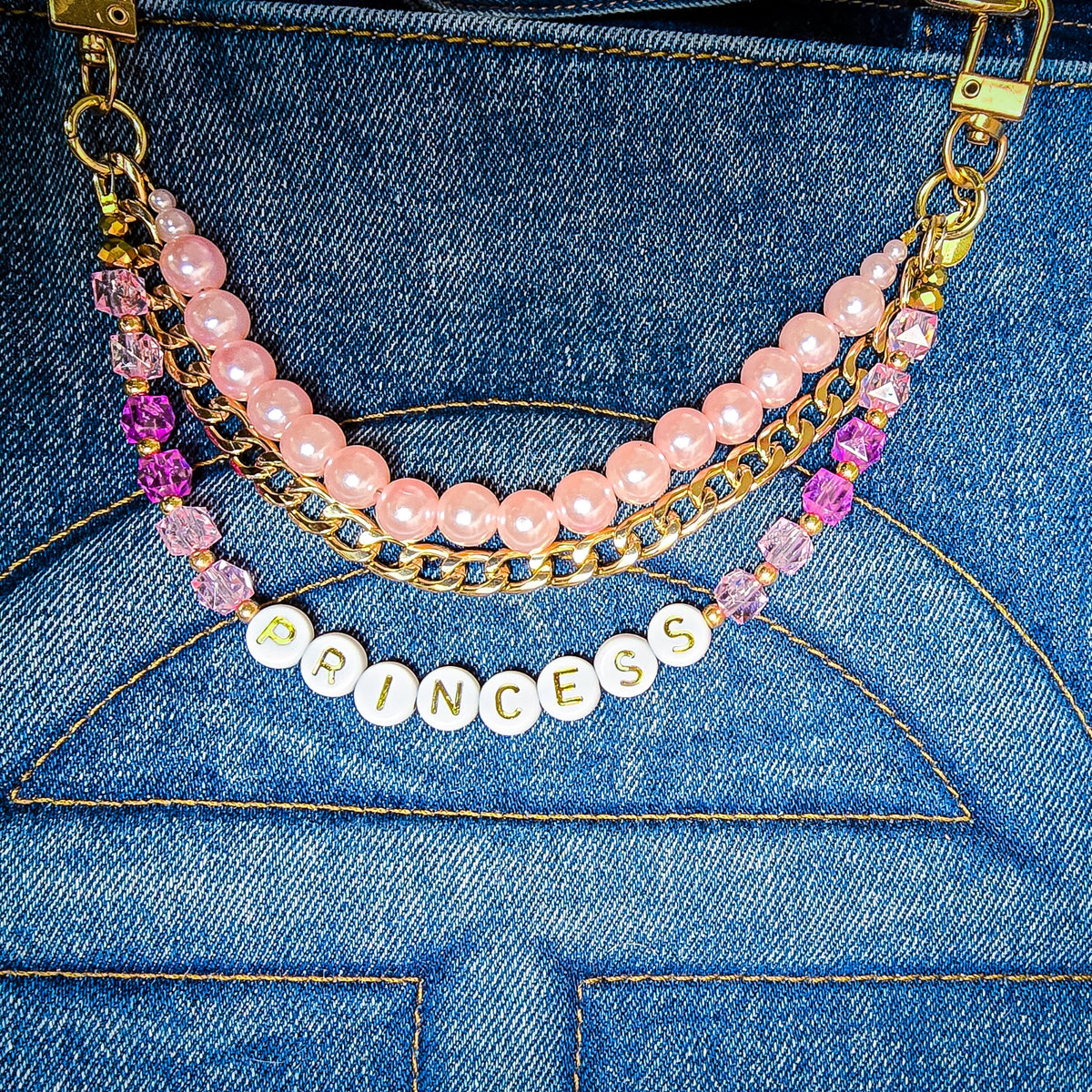 A close-up of a double-layered beaded bag charm draped over a blue denim tote bag. The bag charm features a top layer of light pink pearls and a bottom layer of gold curb linked chains, accented with faceted pink and deep pink crystals and a white round letter bead spelling 'PRINCESS'. All hardware is gold including the lobster clasp closures.