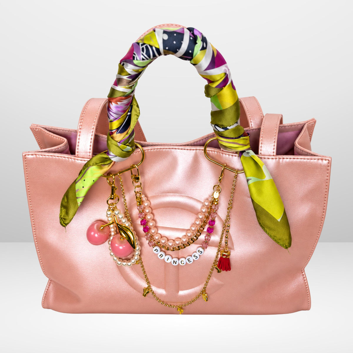 A ballerina pink satin textured tote bag with a colorful patterned scarf wrapped around its handles, adorned with gold-tone & pink charms, set against a light gray background.