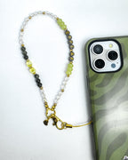 A beaded phone strap with gold-toned hardware and a small bell, shown next to a smartphone on a white background. The beads include clear faceted stones, dark gray beads, light green beads, and yellow-green translucent beads, with some beads spelling out a name.