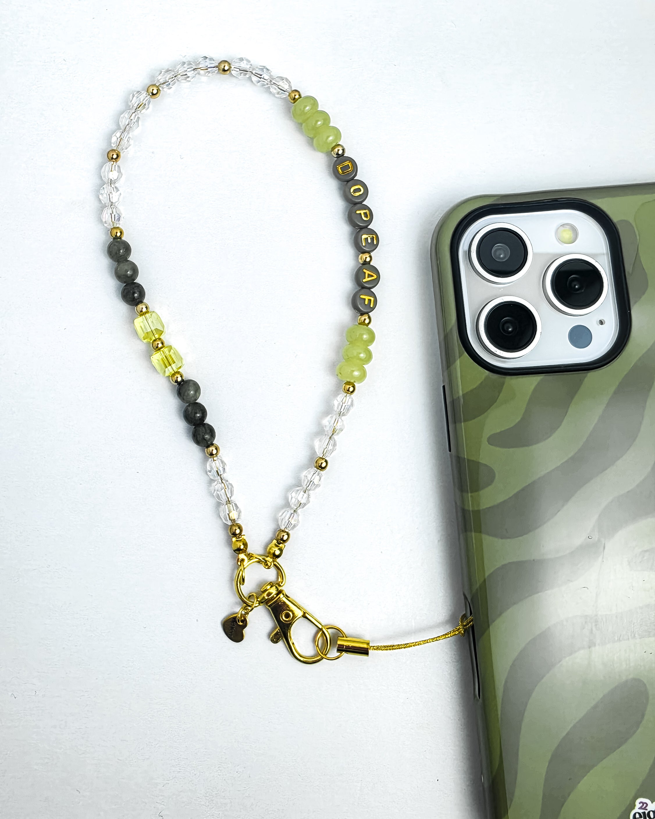 A beaded phone strap with gold-toned hardware and a small bell, shown next to a smartphone on a white background. The beads include clear faceted stones, dark gray beads, light green beads, and yellow-green translucent beads, with some beads spelling out a name.