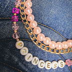 A detailed close-up of a double-layered beaded bag charm draped over a blue denim tote bag. The bag charm features a top layer of light pink pearls and a bottom layer of gold curb linked chains, accented with faceted pink and deep pink crystals and a white round letter bead spelling 'PRINCESS'. All hardware is gold including the lobster clasp closures.