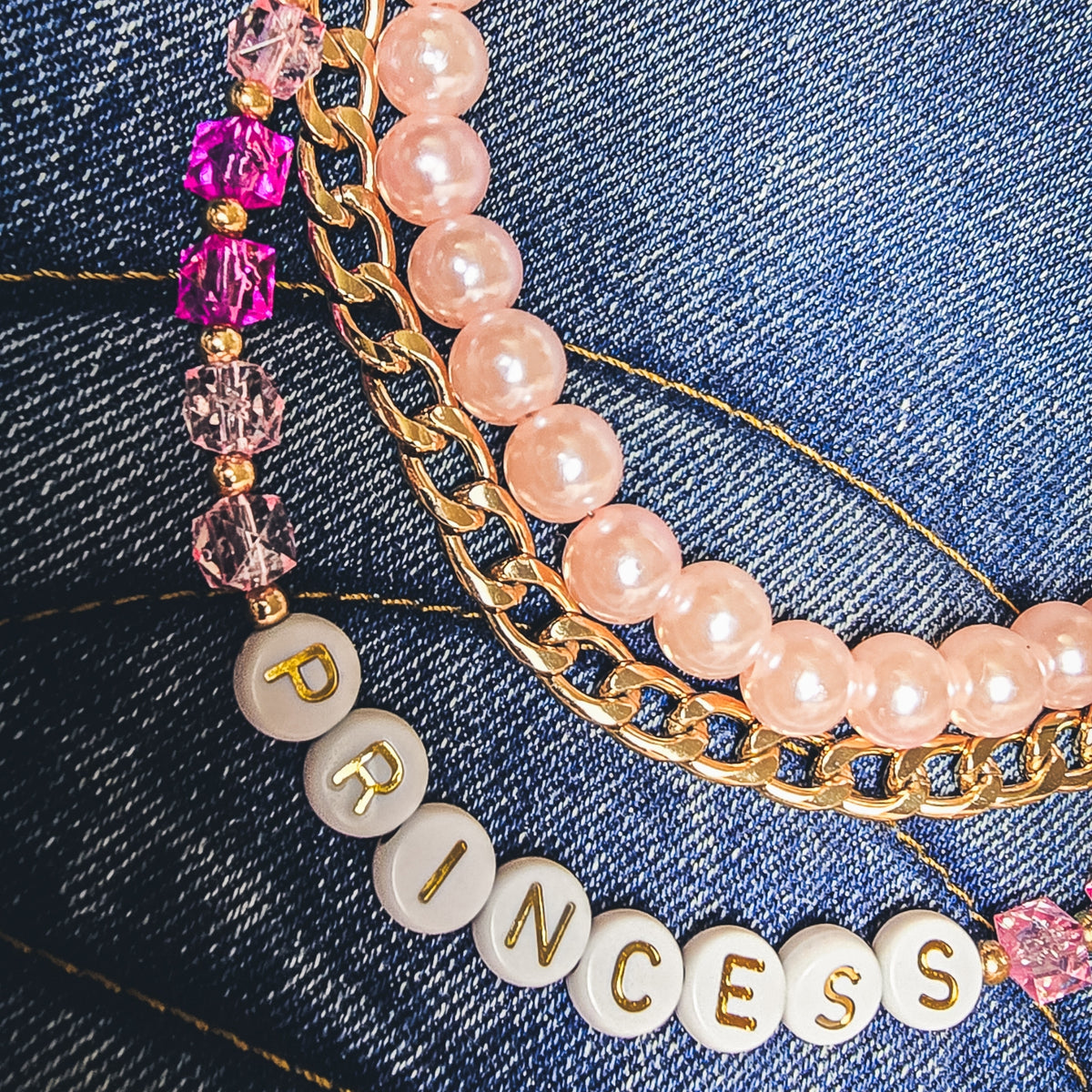 A detailed close-up of a double-layered beaded bag charm draped over a blue denim tote bag. The bag charm features a top layer of light pink pearls and a bottom layer of gold curb linked chains, accented with faceted pink and deep pink crystals and a white round letter bead spelling 'PRINCESS'. All hardware is gold including the lobster clasp closures.