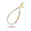 A gold and fine crystal phone charm with amber and lavender beaded accents. Round, white beads spell out "BLESSED" in gold letters on the charm. Accented with a heart shaped brand tag attached to the golden lobster claw clasp