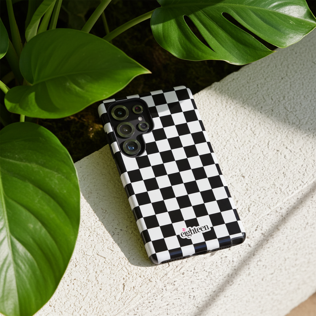 Black and white checkered phone case with 'eighteen' branding on a light surface with green leaves.