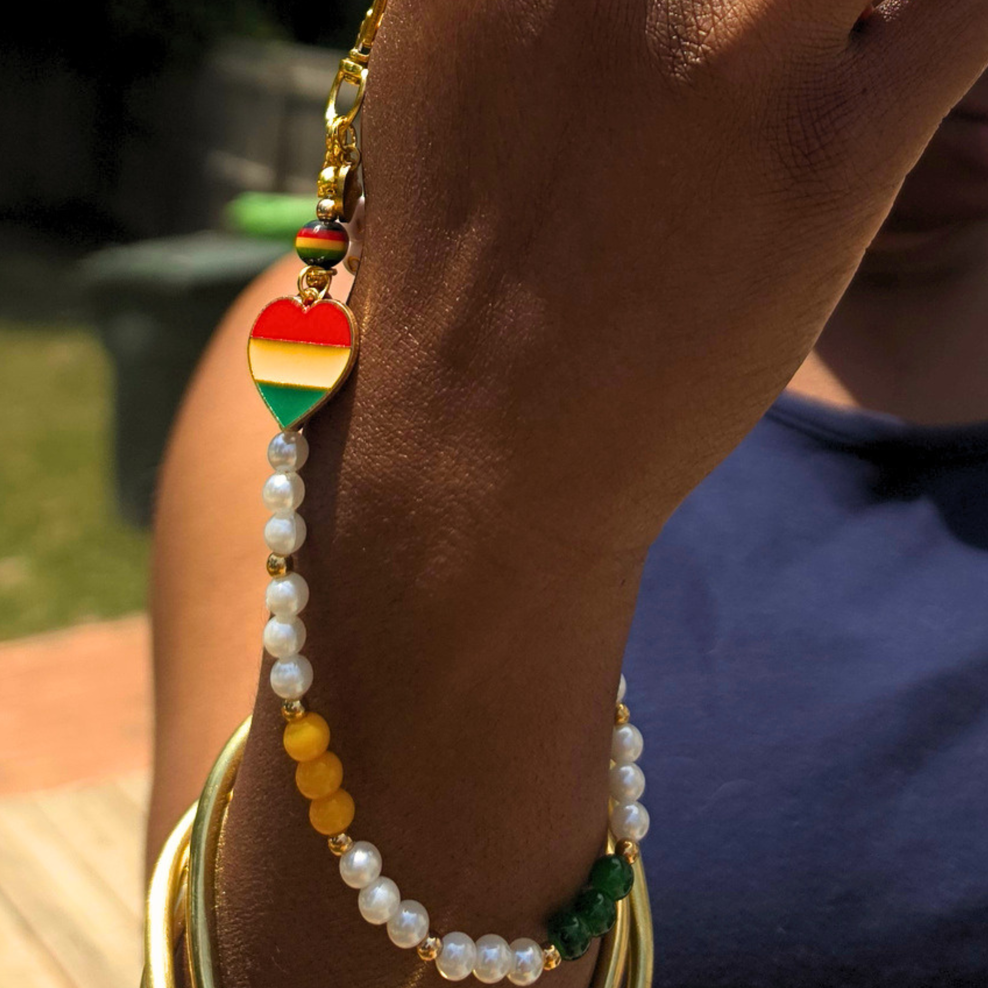A close-up of a person's wrist wearing a beaded bracelet with a heart-shaped Rastafarian flag charm, white pearls, yellow beads, green beads, and gold-toned metal.