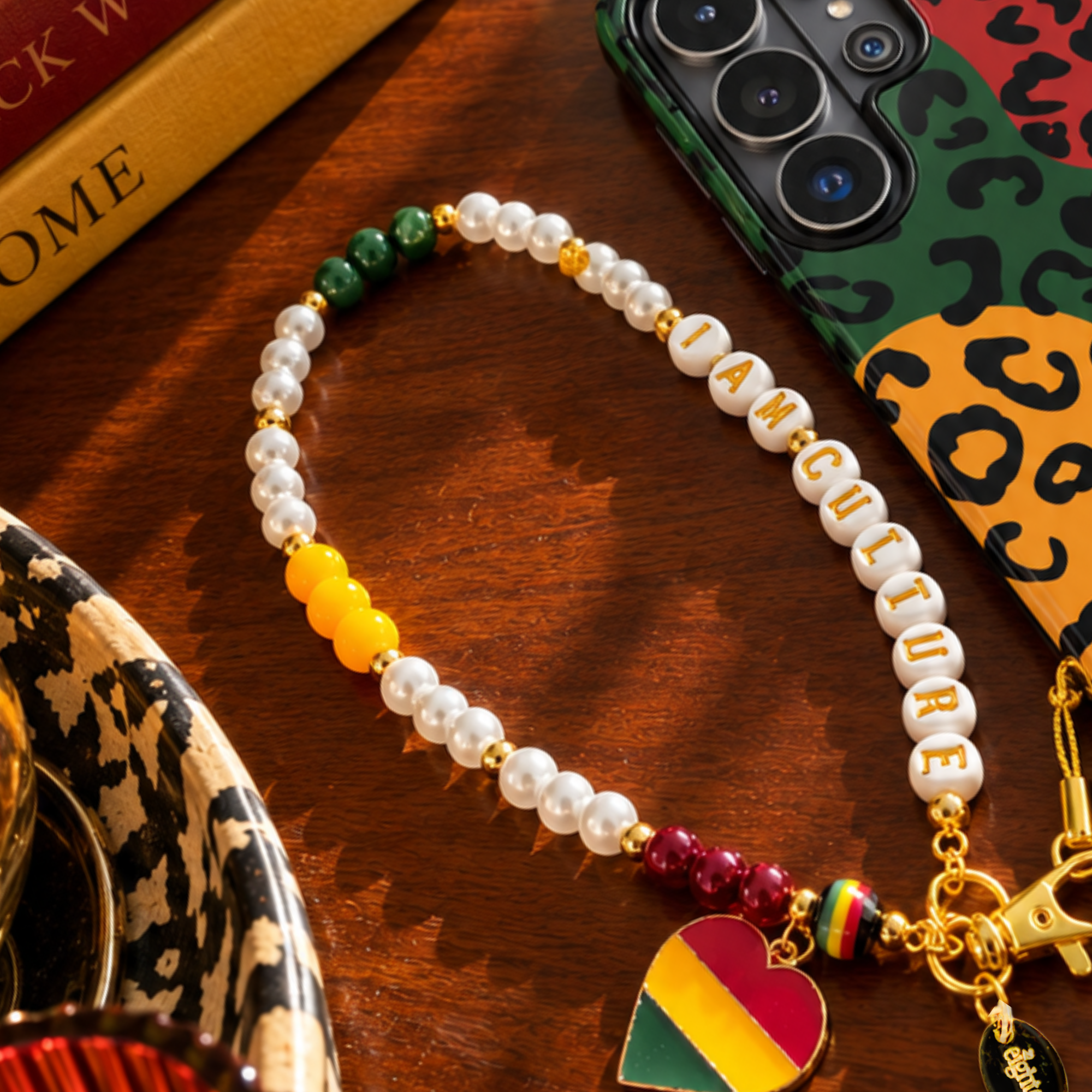 A beaded bracelet with white pearl-like beads, green beads, yellow accent beads, and letter beads spelling 'I AM CULTURE', displayed on a wooden surface next to a leopard-print phone case and books.